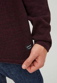 Pull en tricot bordeaux avec un motif texturé, doté d'une étiquette de marque noire sur l'ourlet inférieur et de manches longues.