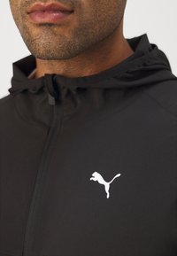 Schwarze Zip-Jacke aus leichtem Material mit einem weißen Puma-Logo auf der linken Brustseite und Kapuze.