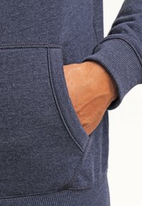 En vuxenhand är delvis inslagen i framfickan på en marinblå sweatshirt med synlig sömnad och ribbad mudd.