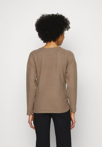 Camisola de manga longa, de tecido canelado em tom castanho claro, com um decote arredondado e corte descontraído, apresentando costuras visíveis na parte de trás.