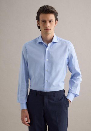 Giovane uomo in piedi con una mano in tasca, indossa una camicia azzurra chiara e pantaloni blu scuro su uno sfondo neutro.