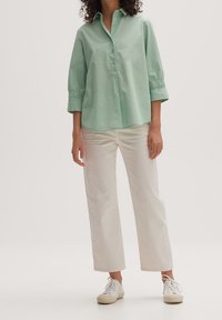 Ljusgrön, långärmad skjorta med krage och veckade manschetter, kombinerad med vita raka byxor och vita sneakers.