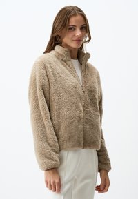 Beige fleece ritsjack met hoge kraag, gestructureerde stof en verlaagde schouders, gedragen over een lichtgekleurde top.