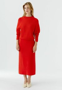 Frau mit blondem Haar, die einen roten Pullover mit langen Ärmeln und einen passenden roten Midirock trägt, kombiniert mit cremefarbenen spitzen Schuhen, steht vor weißem Hintergrund.