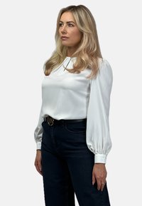 Biała satynowa bluzka z bufiastymi długimi rękawami, z niewielkim detalem dekoltu, w połączeniu z ciemnoniebieskimi dżinsami i czarnym paskiem z okrągłą klamrą.