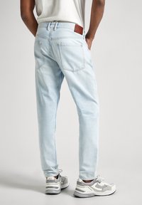 Persona vestida con jeans azul claro y zapatillas blancas con detalles grises, de pie con las manos en los bolsillos frente a un fondo liso.