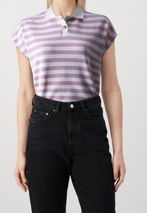 Vrouw draagt een kortärmelig polohemd met lavendel- en wit gestreept patroon, ingestopt in een zwarte, hoog getailleerde jeans, staand tegen een effen achtergrond.