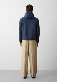 Giacca impermeabile blu navy con cappuccio alto e strutturato, orlo elasticizzato e due tasche laterali, abbinata a pantaloni beige chiaro a gamba larga.