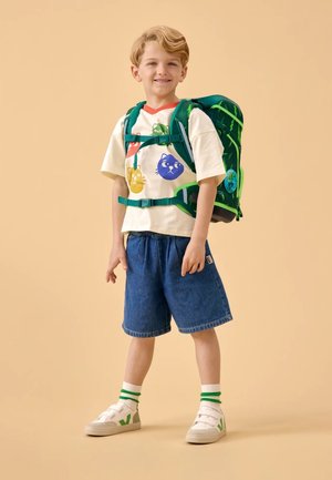 Groene rugzak met verschillende zakken, gecombineerd met een crème T-shirt met kleurrijke diergrafieken. Blauwe denim shorts en witte sneakers.