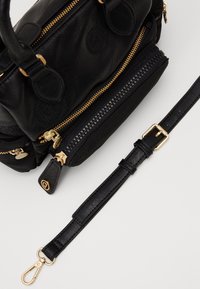 Zwarte leren handtas met ritsen en hardware in goudkleur, voorzien van dubbele bovenhandvatten, een afneembare riem en meerdere met ritssluitingen afgesloten vakken.