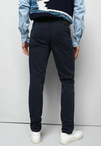 Homme portant un pantalon bleu foncé, une chemise bleu clair, un gilet à motifs bleu marine et des baskets blanches, se tenant devant un fond gris clair uni.