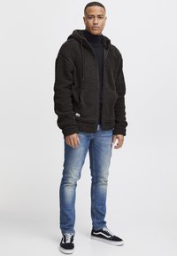 Man wearing a black fleece zip-up hoodie, black turtleneck, blue jeans, and black-and-white sneakers standing against plain background.