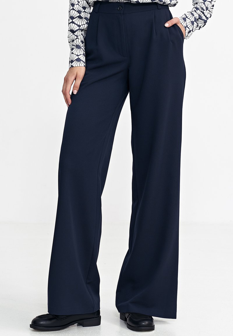 Granatowe spodnie z szerokimi nogawkami, wykonane z gładkiej tkaniny, z wysokim stanem, plisami z przodu i bocznymi kieszeniami. Noszone z czarnymi butami.
