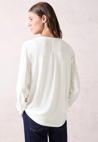 Witte blouse met lange mouwen en een ronde halslijn, subtiele plooien aan de achterzijde en gestructureerde mouwen, gecombineerd met donkerblauwe jeans. Zacht, soepel materiaal.