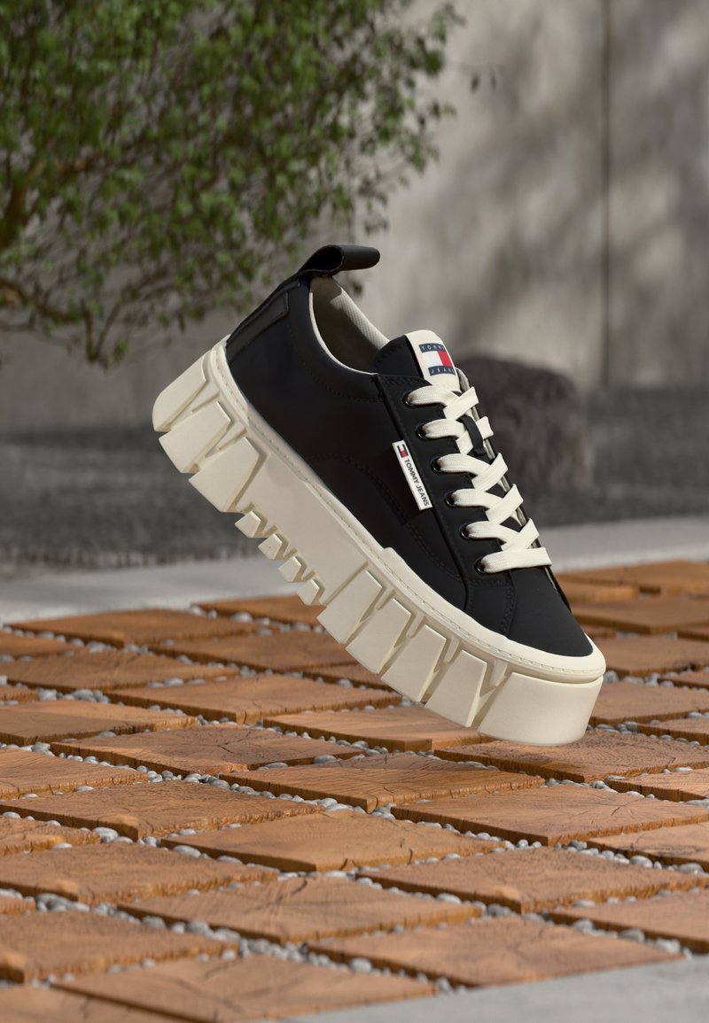 Μαύρο αθλητικό παπούτσι με χοντρή λευκή σόλα και κορδόνια, ετικέτα Tommy Jeans στο πλάι, αιωρούμενο πάνω από ξύλινο πλακάκι σε εξωτερικό χώρο.