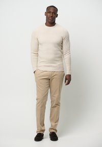 Beige Strickpullover, figurbetont, lange Ärmel. Helle beigefarbene Hose, gerade Beinform, weiche Textur. Dunkelbraune WildlederSlipper, einfaches Design.