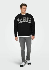 Pull noir avec "PARIS" en grandes lettres blanches, assorti à un jean gris et des baskets noires et blanches. Design simple et décontracté.