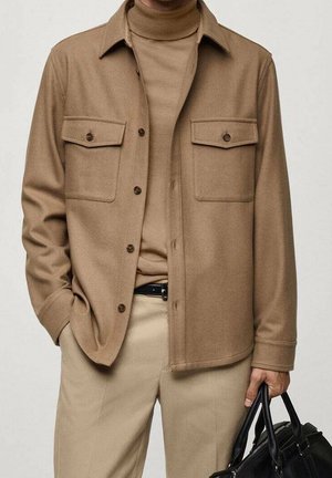 Mann trägt eine hellbraune Hemdjacke über einem passenden Rollkragenpullover und beigen Hosen, hält eine schwarze Lederhandtasche in der rechten Hand.