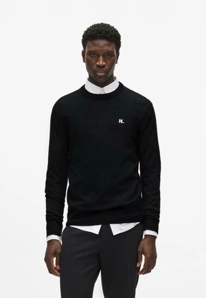 Mann trägt schwarzen Pullover mit langen Ärmeln und weißem Logo, darunter ein weißes Hemd mit Kragen, sowie schwarze Hose, steht vor einem einfarbigen weißen Hintergrund.