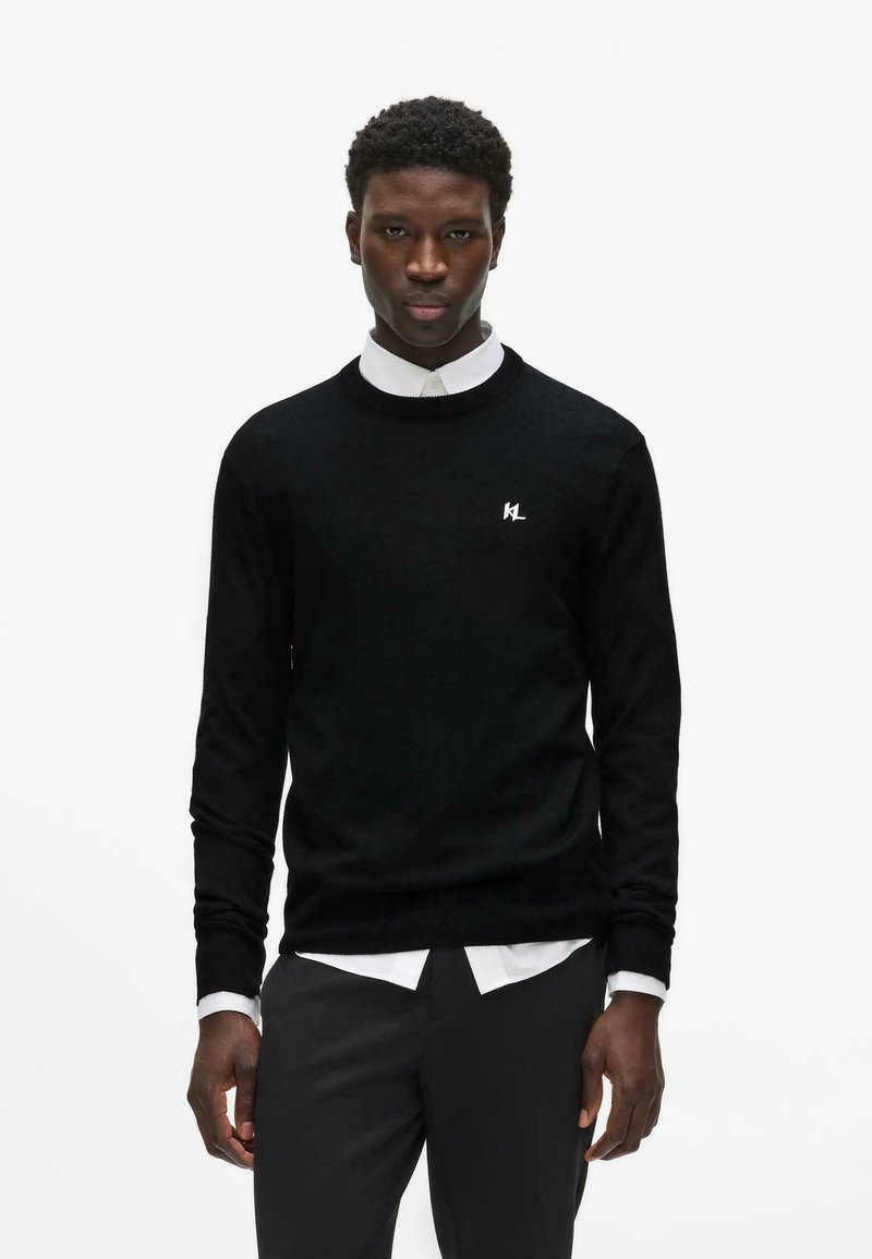 Homme portant un pull noir à manches longues avec un logo blanc, une chemise blanche à col en dessous, et un pantalon noir, debout sur un fond blanc uni.