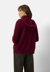 Burgundowy sweter z kapturem o teksturowanym wykończeniu, z wysokim kołnierzem i prążkowanym dołem, zestawiony z ciemnoszarymi spodniami.