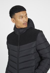 Chaqueta de plumas negra con un panel de forro polar texturizado negro en los hombros, cuenta con capucha y cierre frontal con cremallera.