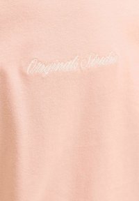 Tissu rose clair avec texte en cursive blanc brodé centré indiquant "Originals Studio".