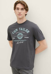 Graues Baumwoll-T-Shirt mit kurzen Ärmeln, mit türkisfarbenem Text, der "Tom Tailor" und "Premium Quality Essentials" enthält. Glatte Textur.