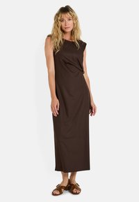 Vestido maxi marrón sin mangas con un corte relajado, que presenta un detalle anudado en la cintura. Combinado con sandalias marrones de tiras.