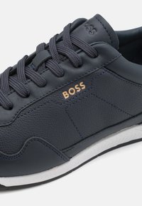 Tamno plave kožne tenisice s teksturiranom površinom, s tamnosivim vezicama, zlatnim logotipom "BOSS" i bijelim gumiranim potplatom s uzorkom za prianjanje.