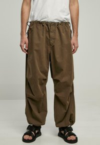 Brune bomuldsbukser med elastisk talje, justerbare snore og en løs pasform. Sammen med sorte sandaler, der har et minimalistisk design.