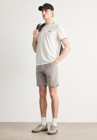 White t-shirt with black trim and logo, light gray shorts, gray shoes, white socks, and a black cap, with a gray jacket draped over one shoulder.