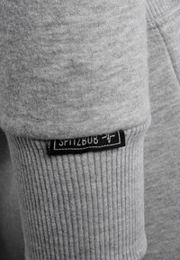 Grauer Sweatshirtstoff mit einer weichen Textur, ausgestattet mit einem gerippten Bündchen und einem schwarzen Etikett mit der Aufschrift "SPITZBUB." Schlichte und funktionale Designdetails.