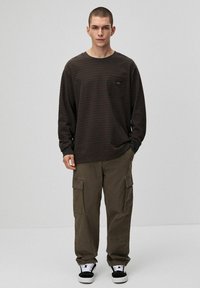 Brązowa koszula w paski z długim rękawem i kieszenią na piersi, zestawiona z luźnymi oliwkowymi spodniami typu cargo oraz czarnymi trampkami z białymi akcentami.