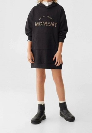 Robe sweat-shirt noire surdimensionnée avec un graphisme "ENJOY EVERY MOMENT" en imprimé floral. Comprend une poche kangourou à l'avant et des poignets côtelés. Portée avec des bottes noires.