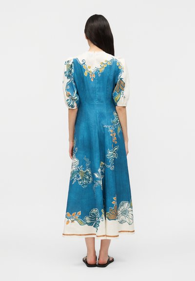 Γυναίκα που φοράει μπλε και άσπρο μίντι φόρεμα με φουσκωτά μανίκια και λουλουδάτα σχέδια, στέκεται ξυπόλυτη, στραμμένη προς το μπροστά, σε απλό φόντο.