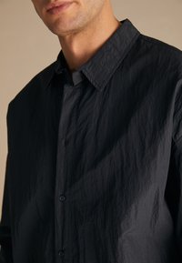 Black, wrinkled fabric shirt with a buttoned front, point collar, and loose design. Features textured material and subtle sheen.