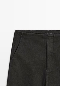 Pantalon en denim noir avec poches latérales, détails de coutures et étiquette de marque visible. Texture lisse avec une coupe ajustée.