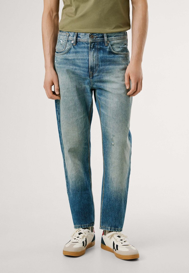 Jeans de mezclilla azul claro con un corte relajado, áreas desgastadas y un ligero distress, combinados con zapatillas blancas y negras.