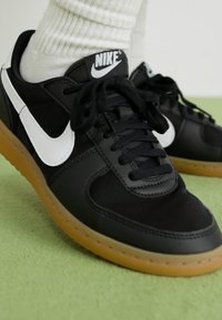 Sorte sneakers med hvid Nike swoosh, med en struktureret overdel og sorte snørebånd, placeret på en gummigummisål mod en grøn baggrund.