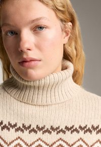 Pulover croșetat bej cu guler înalt, având o textură ribbed și modele geometrice maro pe piept. Vedere detaliată a părții superioare a corpului.