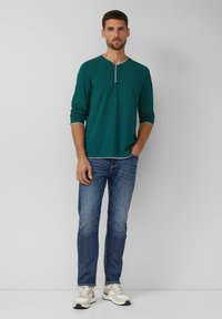 Tannengrünes Langarm-Henley-Shirt mit Kontrastkragen, zwei-Knopf-Leiste, kombiniert mit blauen Jeans und hellen Sneakers.