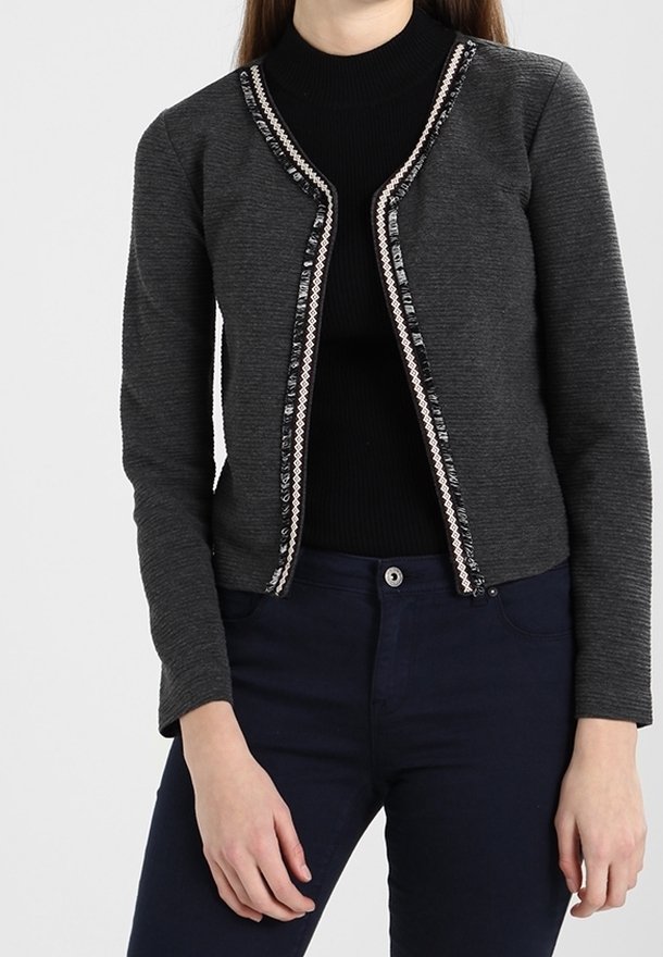 Femme portant un cardigan gris foncé à manches longues et à ouverture frontale avec une bordure décorative, par-dessus un col roulé noir et un pantalon bleu marine.