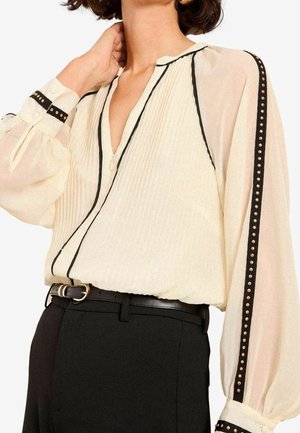 Femme portant un chemisier crème avec un passepoil noir et des rayures cloutées sur les manches, assorti à un pantalon noir taille haute et une ceinture noire.