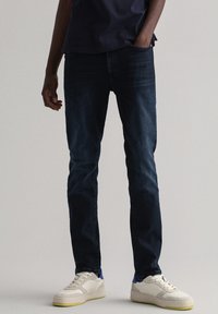 Mørkeblå denimjeans med smal passform, med et klassisk fem-lommers design. Brukt med hvite sneakers med blå og gule aksenter.