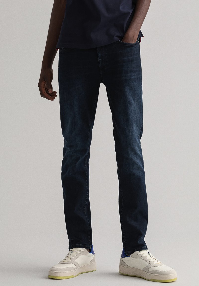 Mørkeblå denimjeans med smal passform, med et klassisk fem-lommers design. Brukt med hvite sneakers med blå og gule aksenter.