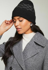 Gorro de punto negro con textura acanalada y detalles de cristales. Combinado con un abrigo de lana gris y un suéter de cuello alto en color crema.