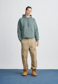 Zelena majica s kapuljačom od flisa s džepom za ruke, bež cargo hlače sa bočnim džepovima i smeđe čizme s vezicama na plavom tepihu.