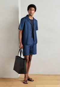 Marineblaues Kurzarmhemd und passende Shorts mit gemustertem Textil, kombiniert mit einer schwarzen Tragetasche und Flip-Flops, vor einem neutralen Hintergrund.