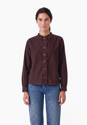 Kvinde med mørkt hår bærer en brun knappet bluse med lange ærmer og flæsedetalje samt blå jeans, stående mod en ensfarvet hvid baggrund.